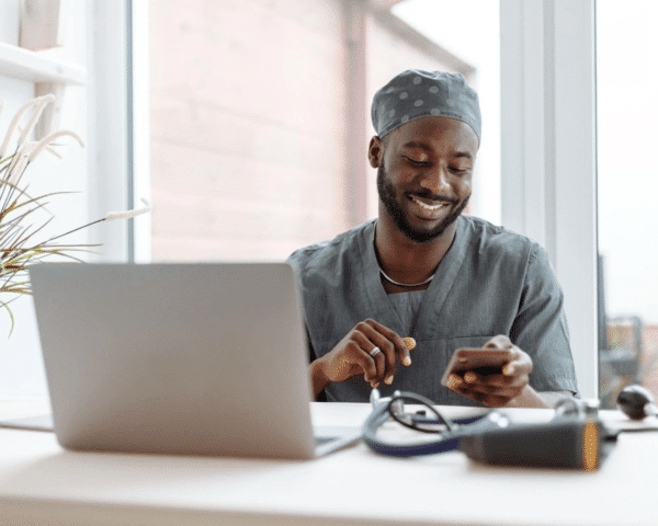 A young doctor smiling and sitting at a desk with his phone and laptop