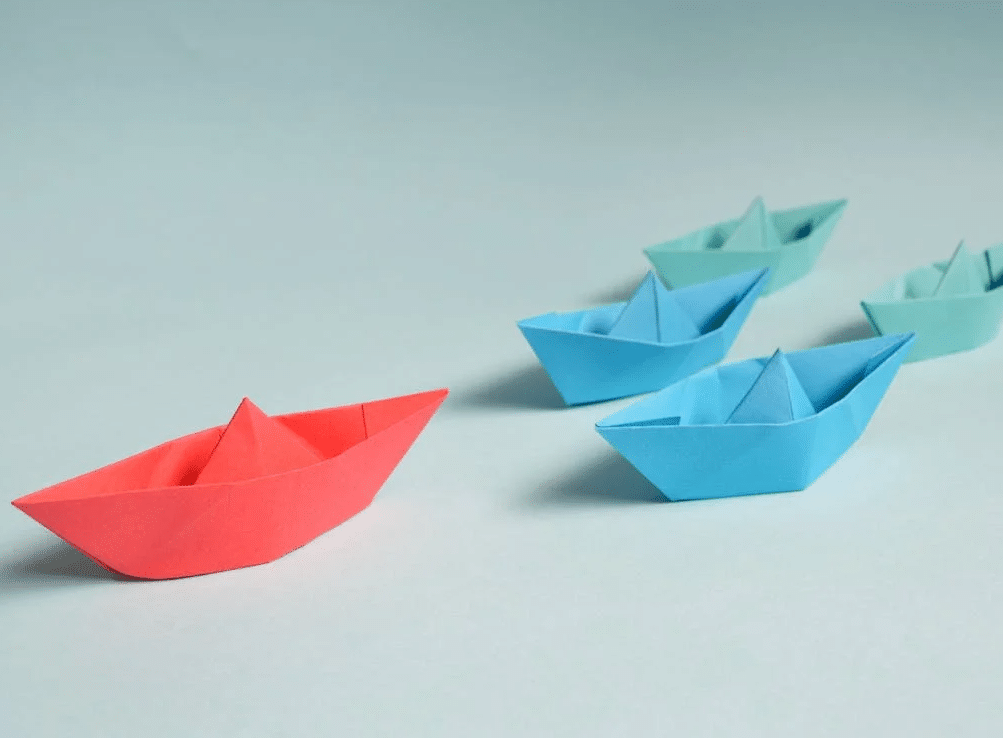 A series of paper boats in different colors, the red one representing the leader
