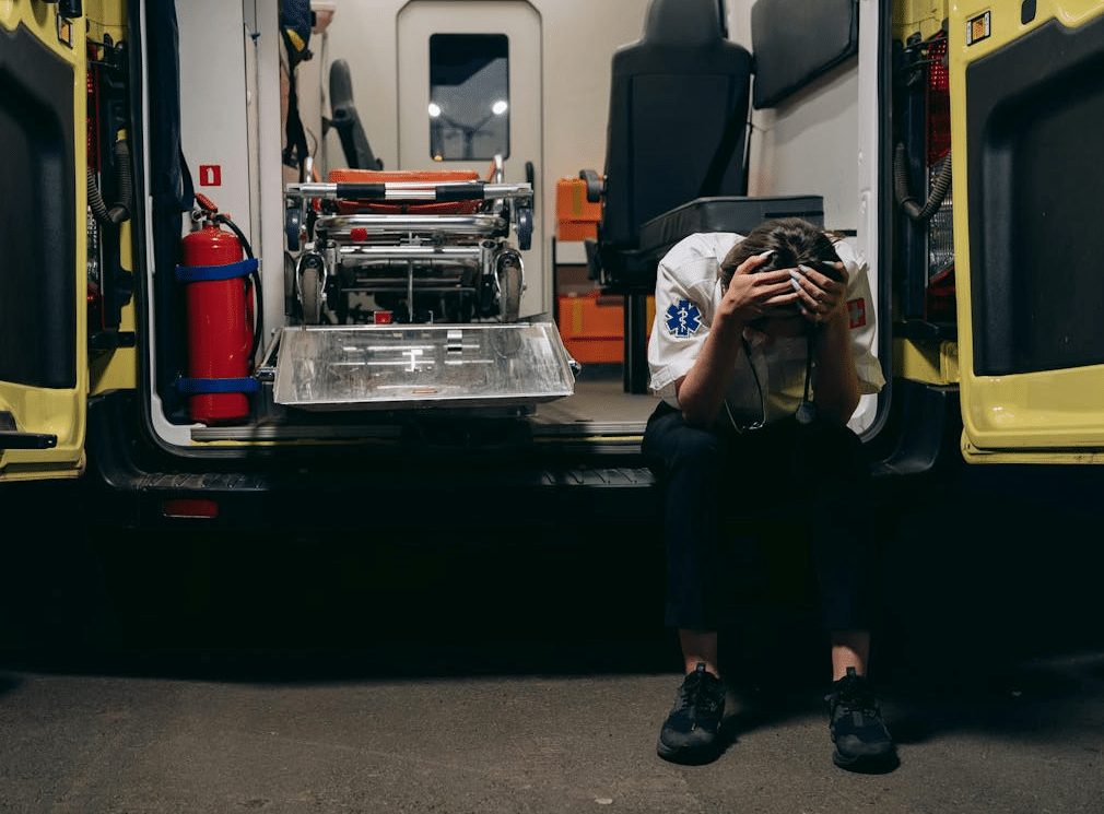 A paramedic sitting on the bumper of an ambulance with head in hands.